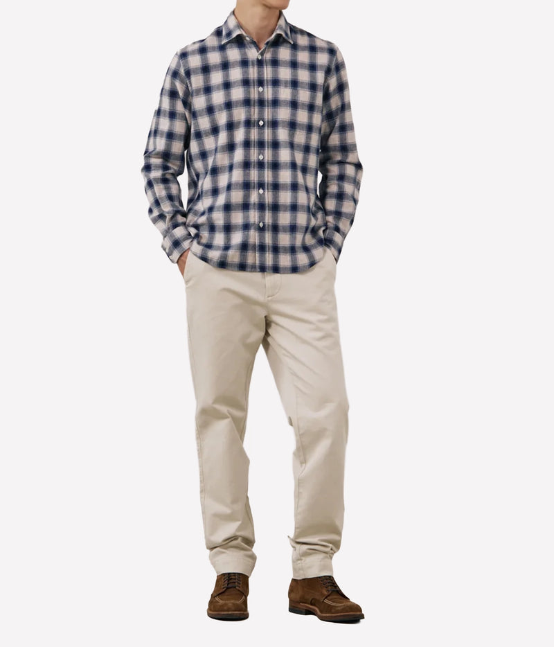 A man wearing a navy and white checkered long-sleeve button-down shirt, beige trousers, and brown shoes, standing against a light grey background.