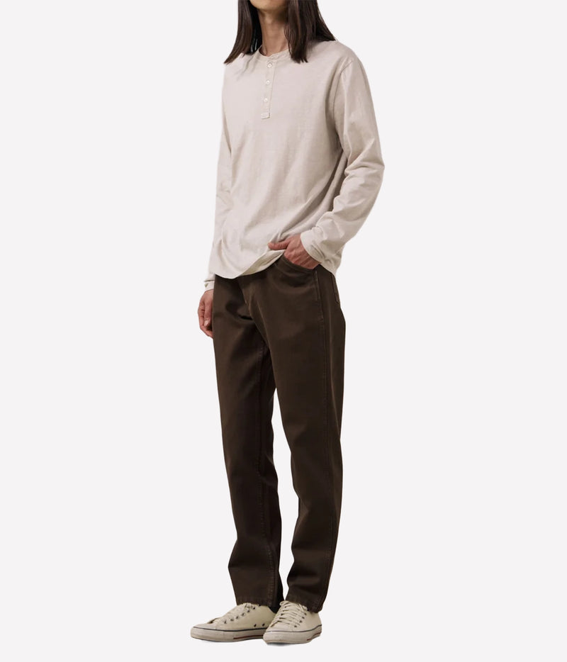A model wearing a light beige long-sleeve henley shirt, dark brown trousers, and beige low-top sneakers, standing against a plain white studio background.