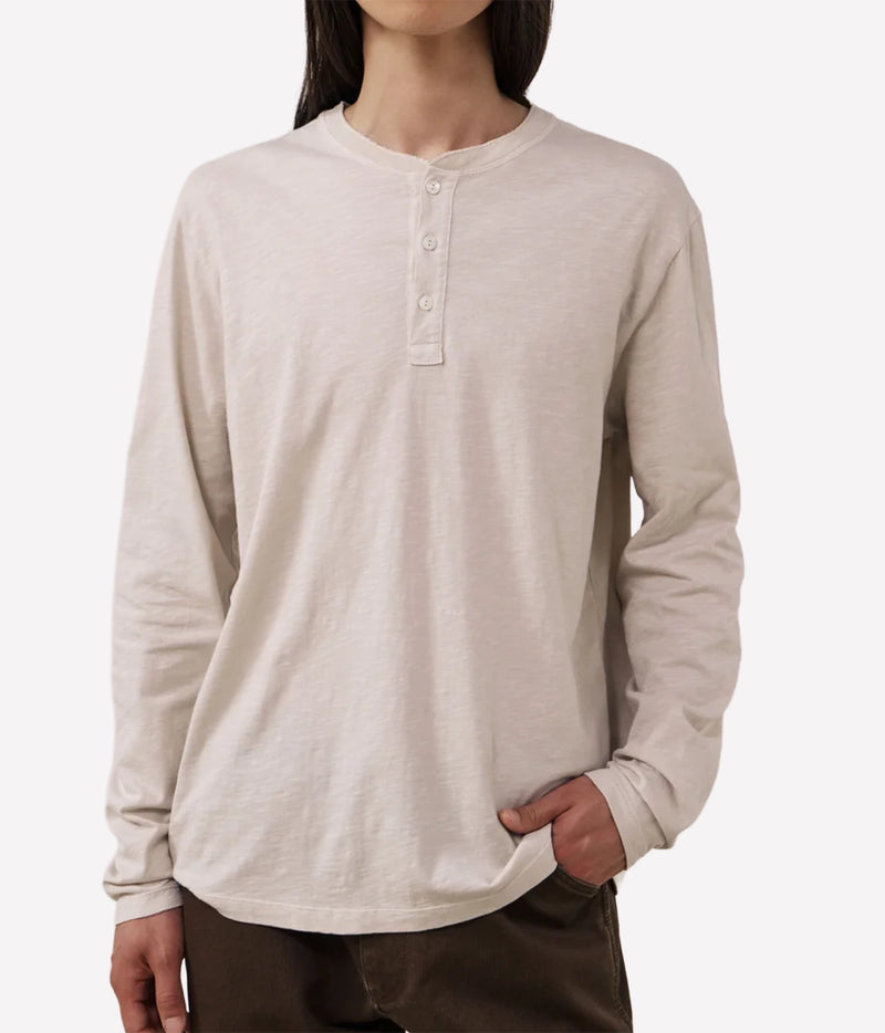 A person wearing a light beige long-sleeve henley shirt with a three-button placket, paired with dark brown pants, against a plain light grey background.