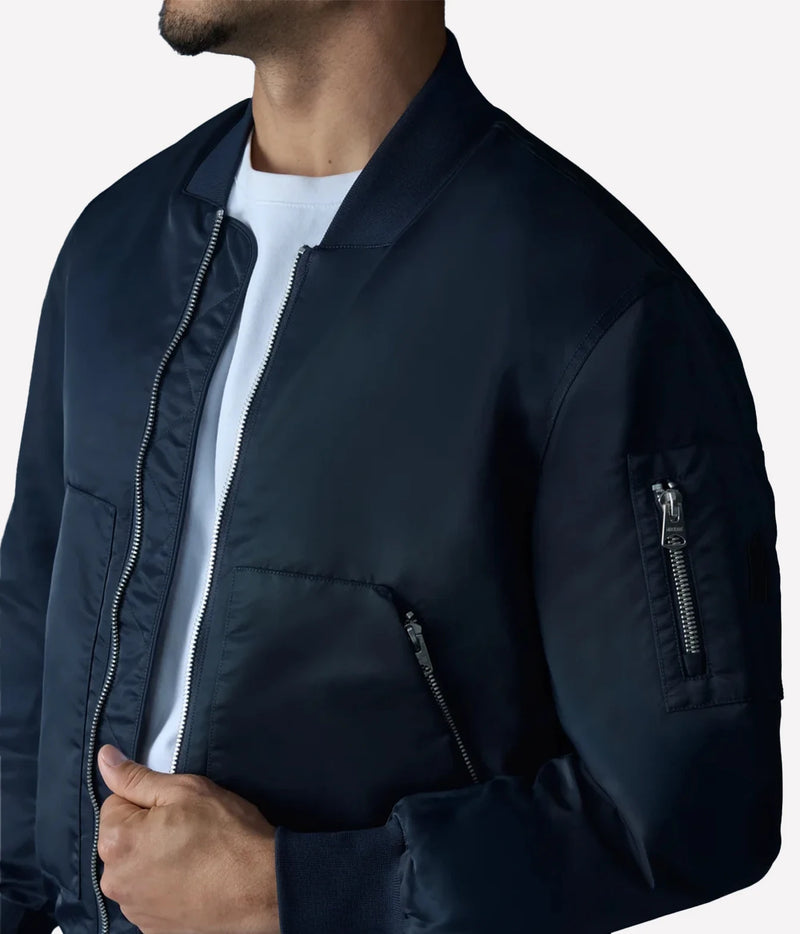 A close-up of a man wearing a dark navy blue bomber jacket over a white t-shirt, showing the chest, collar, and sleeve details against a light gray background.