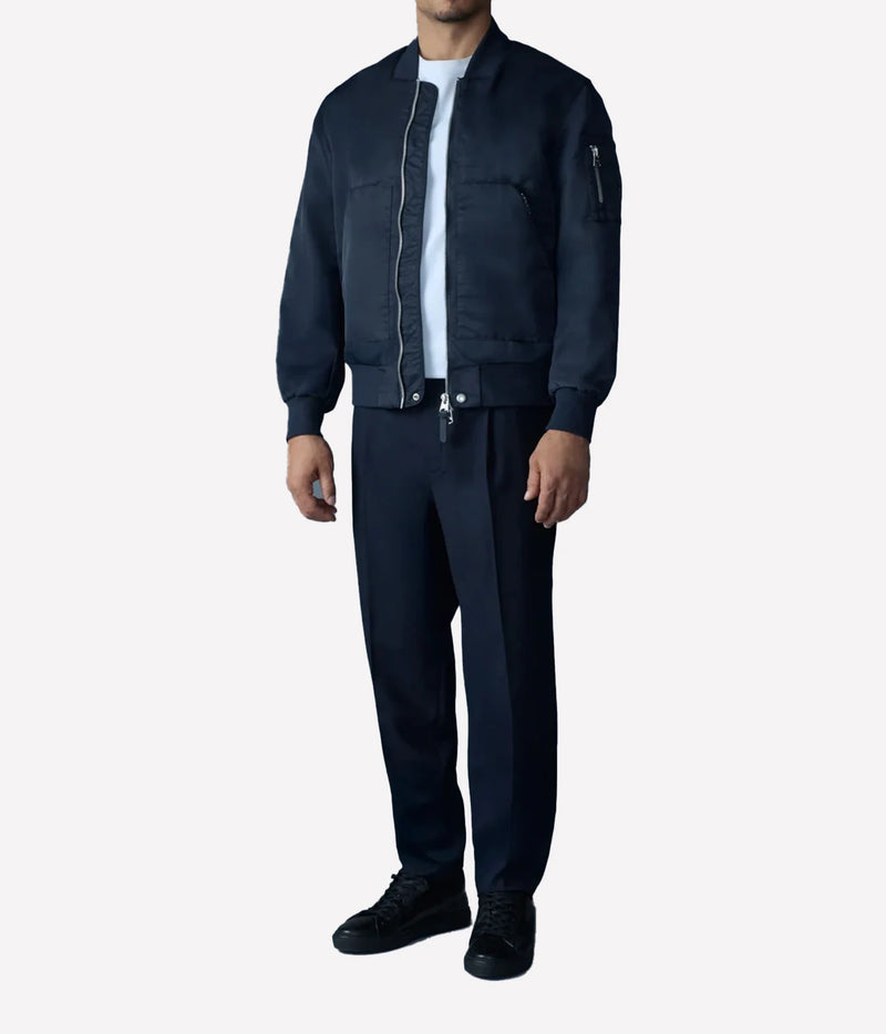 A man wearing a dark navy blue bomber jacket, matching navy trousers, a white t-shirt, and black sneakers, standing against a light gray background.
