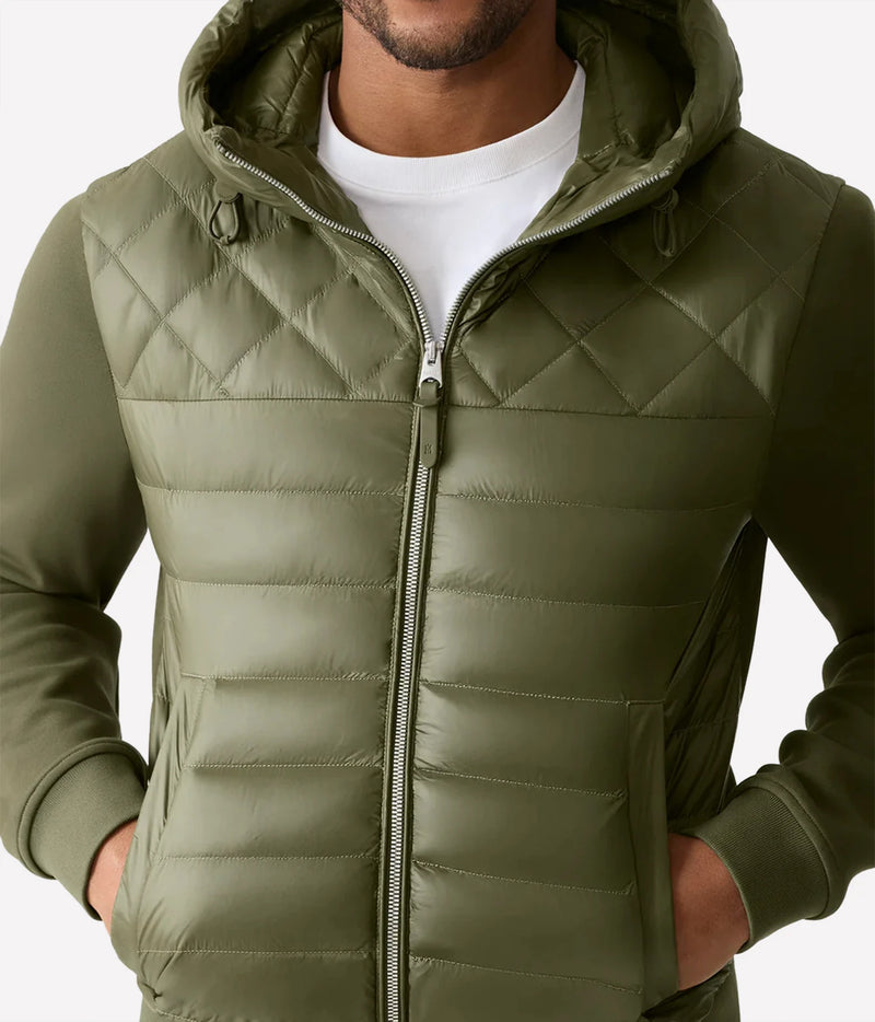 An olive green hooded puffer jacket with diamond quilting, worn by a model over a white t-shirt against a light gray background.