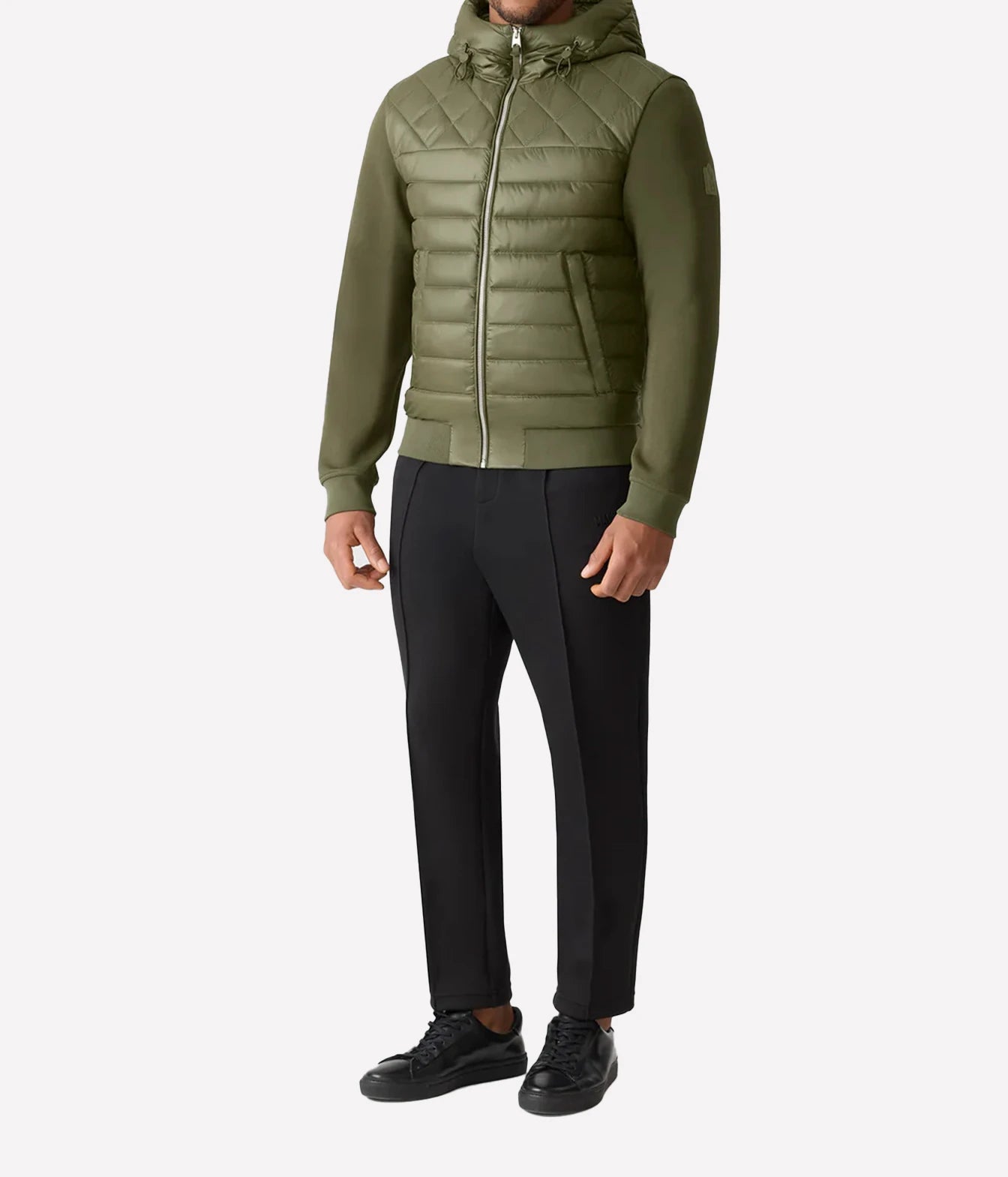 A man wearing an olive green hooded puffer jacket with black trousers and black shoes, standing against a light gray studio background.