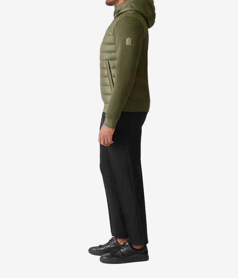 A side profile view of a man wearing an olive green hooded puffer jacket, black trousers, and black sneakers against a light gray background.