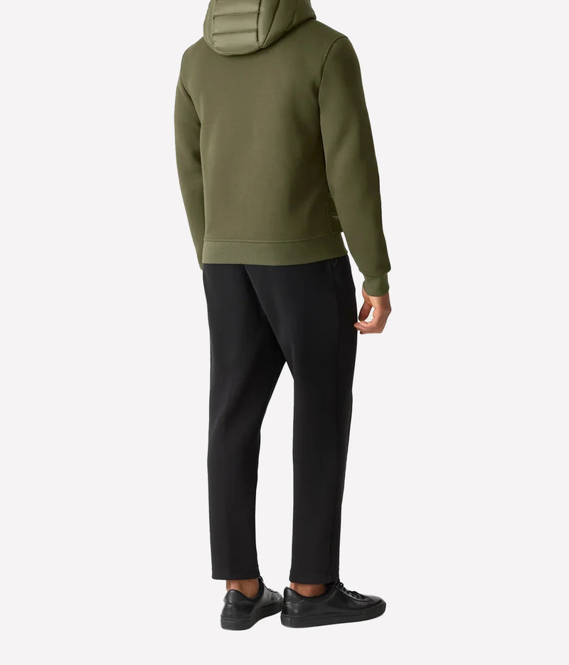 A person shown from the back wearing an olive green hooded sweatshirt, black pants, and black sneakers against a plain light gray studio background.