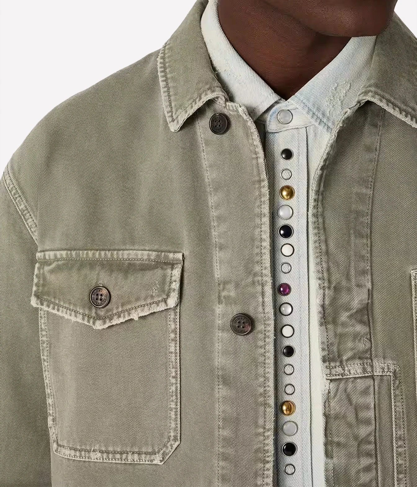 Close-up of a person wearing a light sage green denim jacket over a white button-up shirt with decorative colorful buttons down the front.
