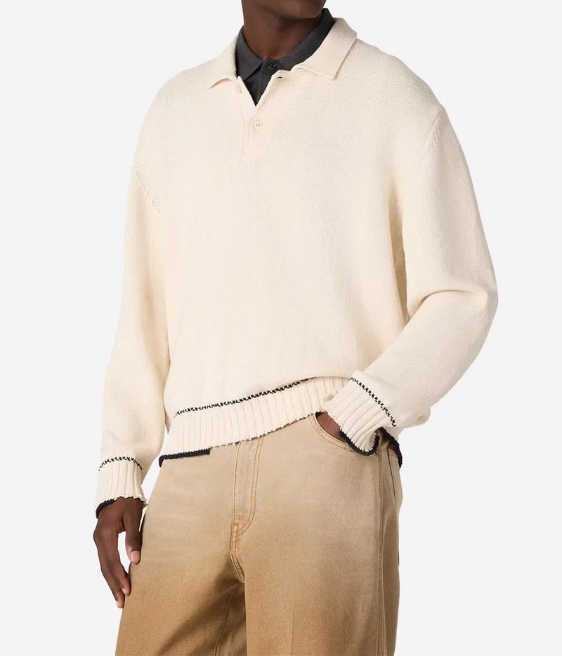 A cream-colored knit polo sweater with black contrast trim, worn by a model over a dark collared shirt and tan trousers, shot against a plain light gray background.