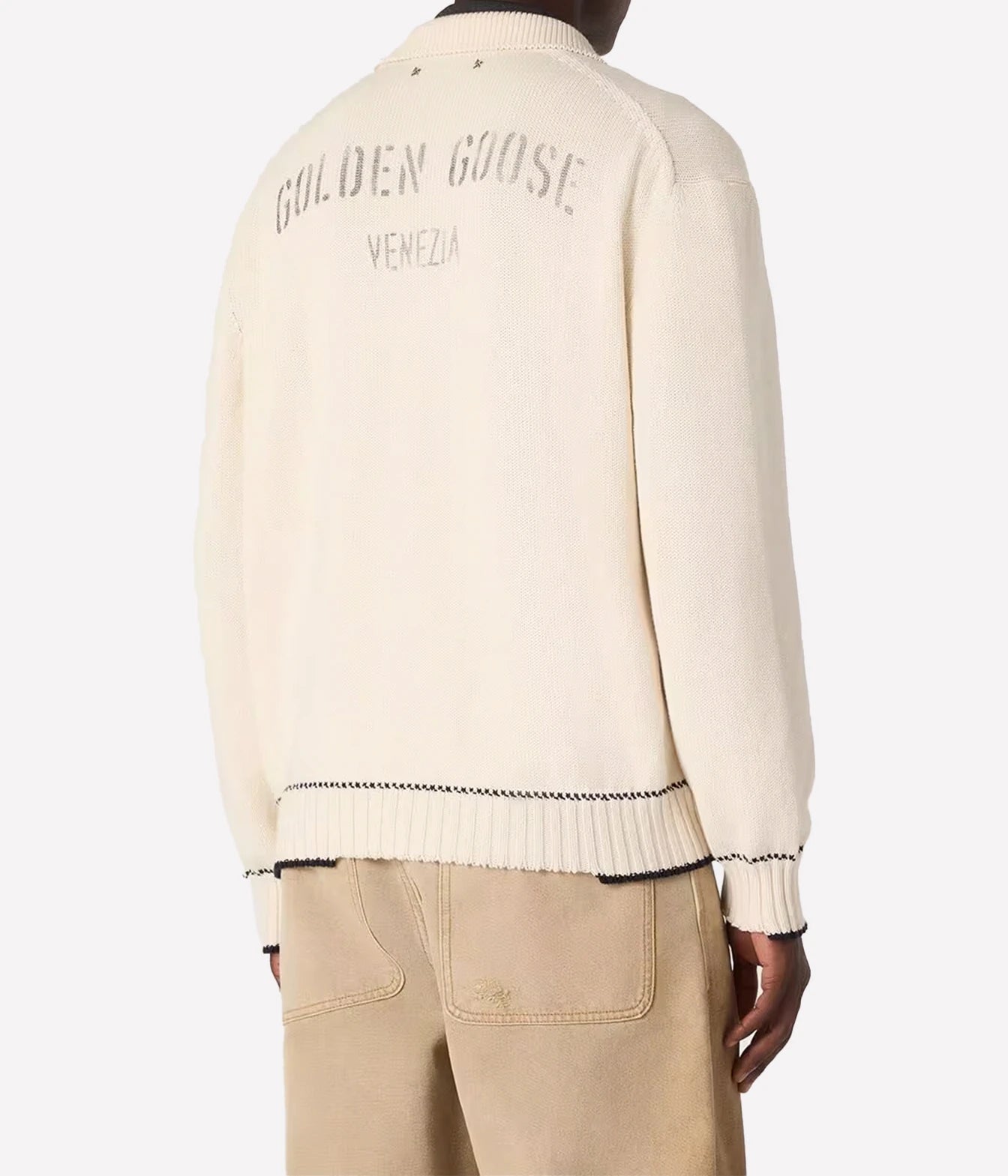 Back view of a cream-colored knit sweater with 'GOLDEN GOOSE VENEZIA' embroidered on the back, worn by a model with beige trousers against a light gray background.