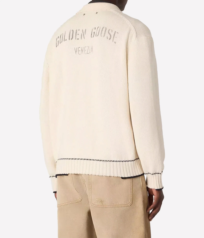 Back view of a cream-colored knit sweater with 'GOLDEN GOOSE VENEZIA' embroidered on the back, worn by a model with beige trousers against a light gray background.
