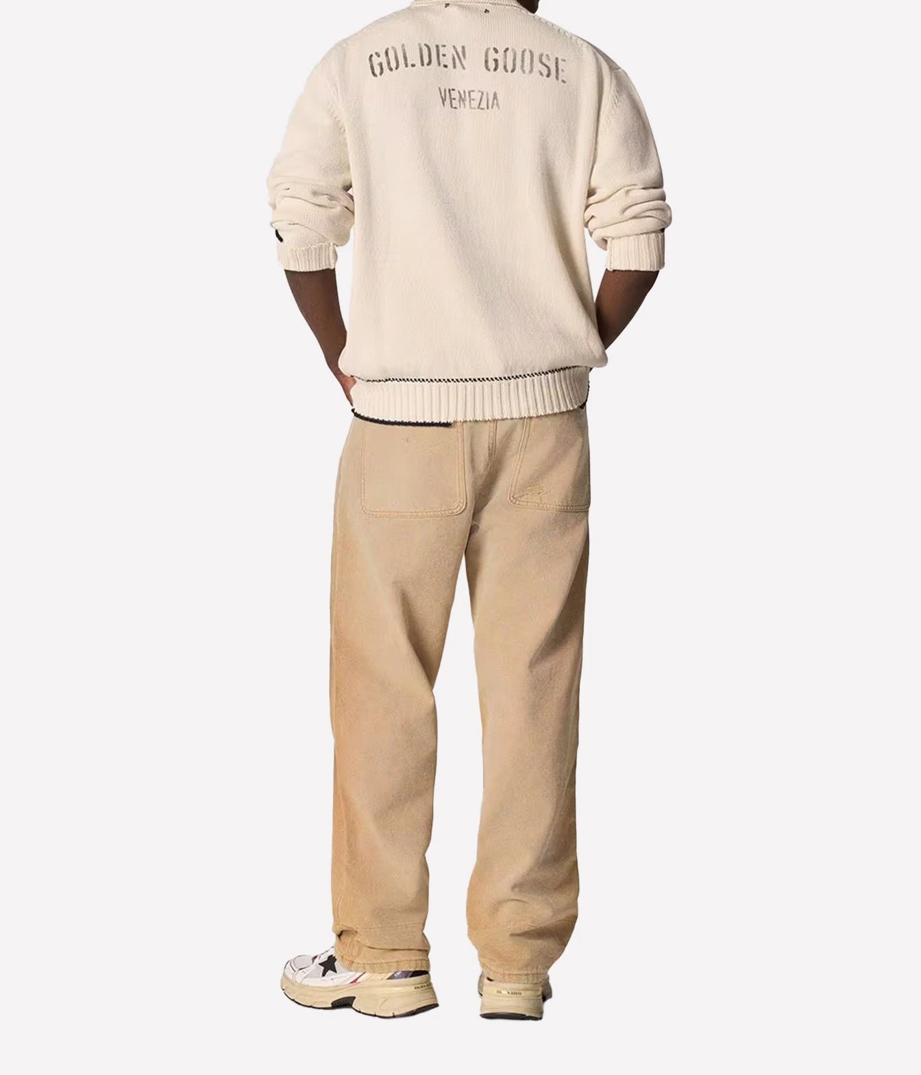 A person shown from the back wearing a cream-colored Golden Goose sweater with 'GOLDEN GOOSE VENEZIA' printed on the back, paired with beige pants and white sneakers, against a light gray background.