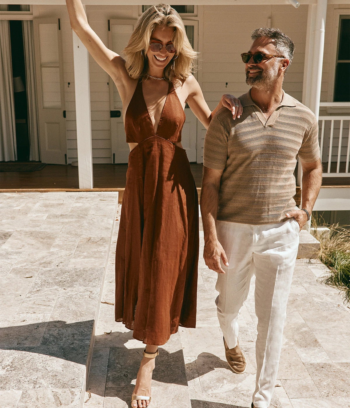 Model wearing the Orlebar Brown Griffon Italian Linen Trouser in White Sands with a light brown striped knit polo, walking alongside a woman in a rust midi dress