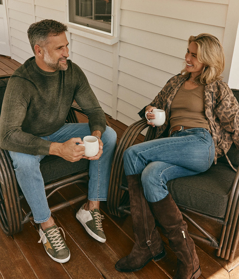 Man wearing Avant Toi Hand Painted Light Cashmere Round Neck Pullover in Moss with jeans, seated on a porch with a coffee