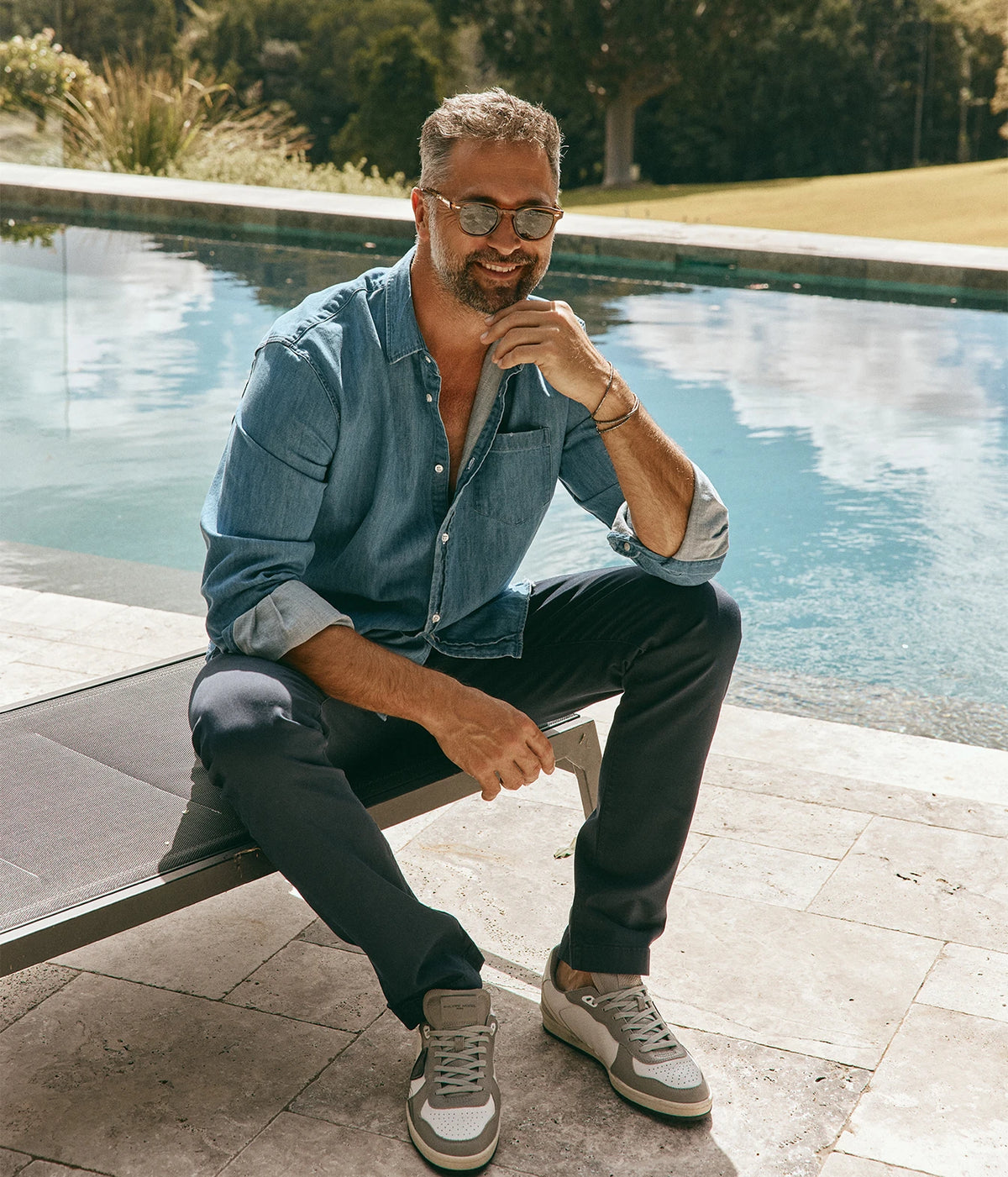 Model wearing the Frank + Eileen Luke Woven Shirt in Stonewashed Indigo Denim with sleeves rolled, dark jeans and sneakers, seated with his arms resting on his knees