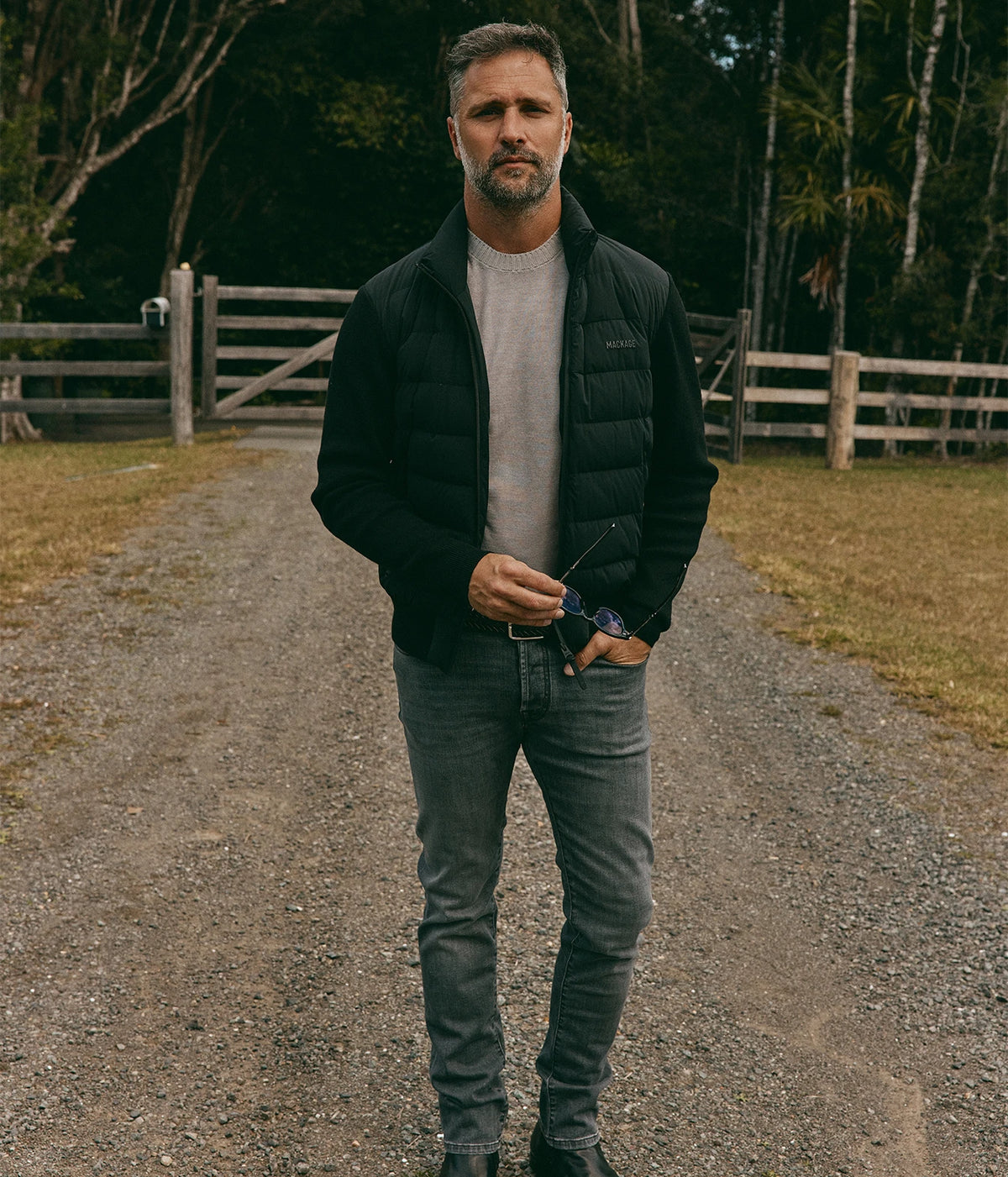 Model wearing the Mackage Haney Jacket in Black open over a grey tee with washed grey jeans, one hand in his pocket