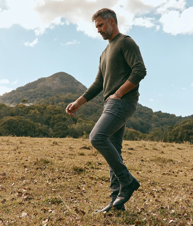 Man wearing Avant Toi Hand Painted Light Cashmere Round Neck Pullover in Moss with jeans, walking through a field