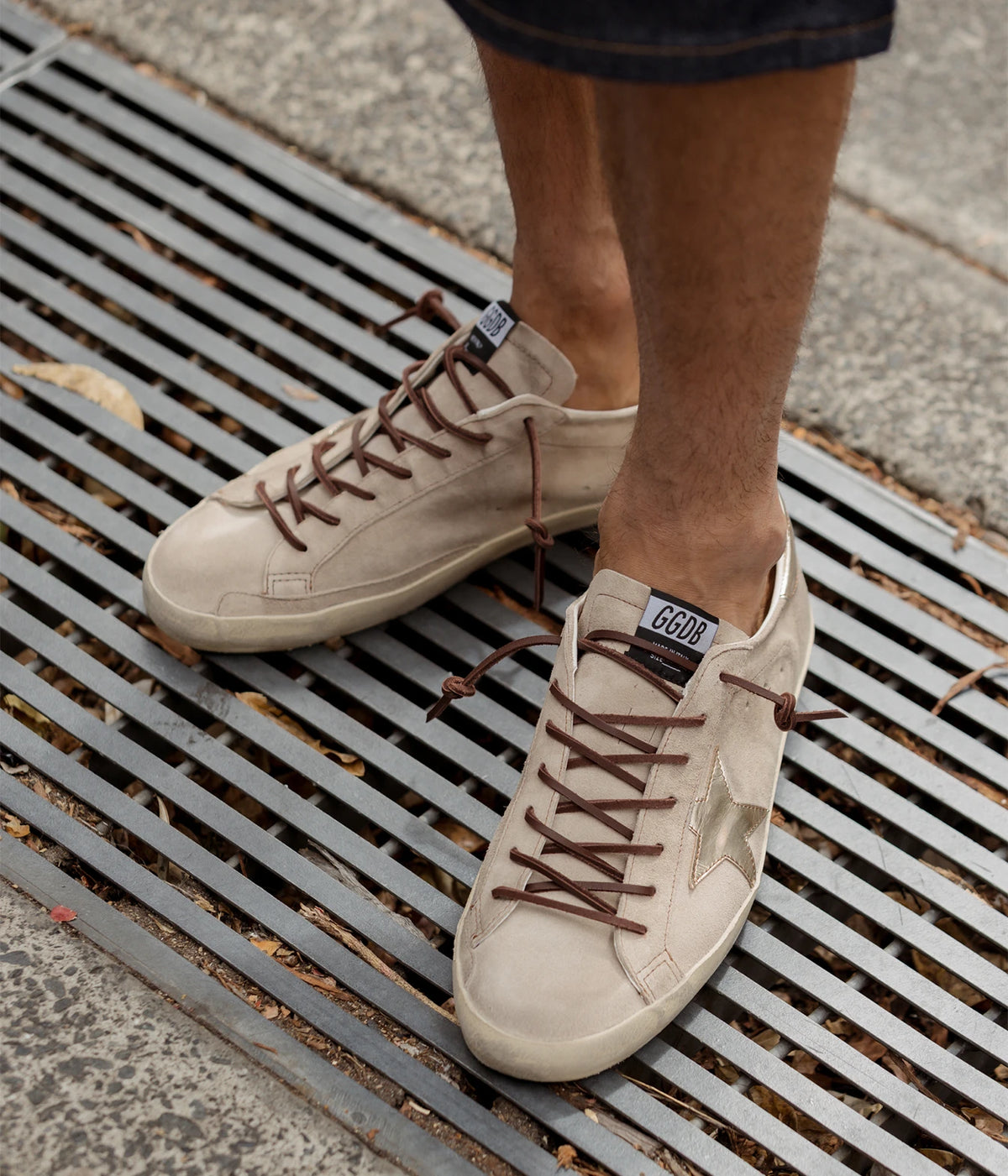 Man wearing Golden Goose Super-Star Sneaker in Beige & Platinum with dark shorts, standing on a metal grate, front view