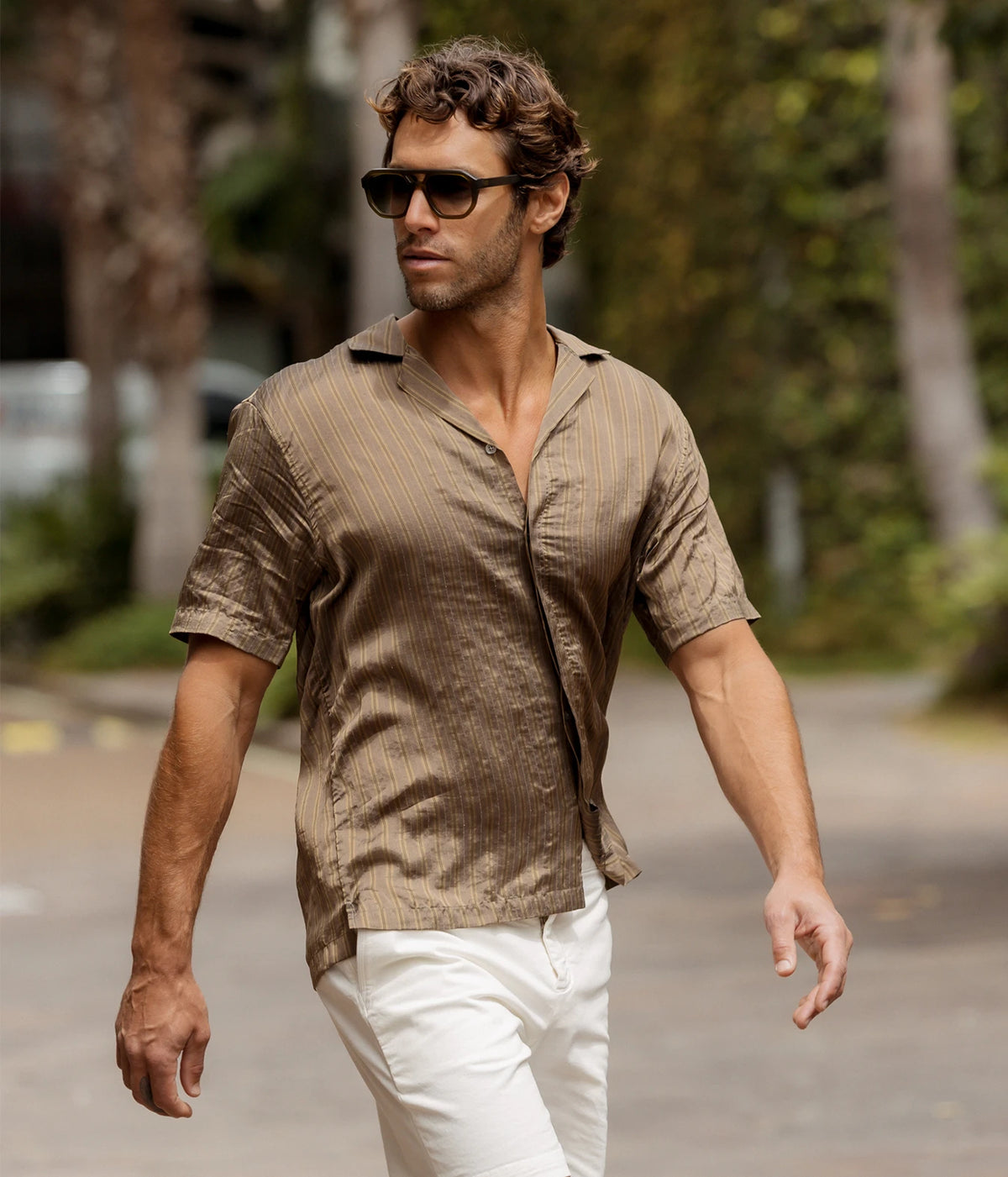Man wearing Orlebar Brown Maitan Villa Stripe Shirt in Bronze with white trousers and sunglasses, walking outdoors, front view
