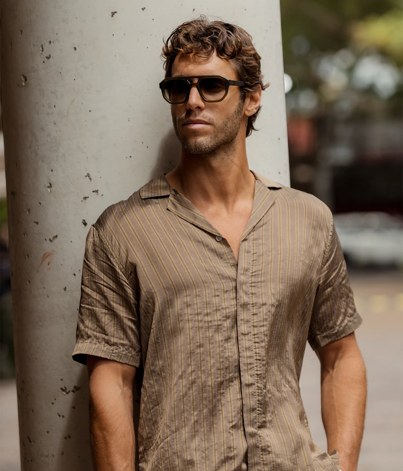 Man wearing Orlebar Brown Maitan Villa Stripe Shirt in Bronze with sunglasses, leaning against a pillar, front view