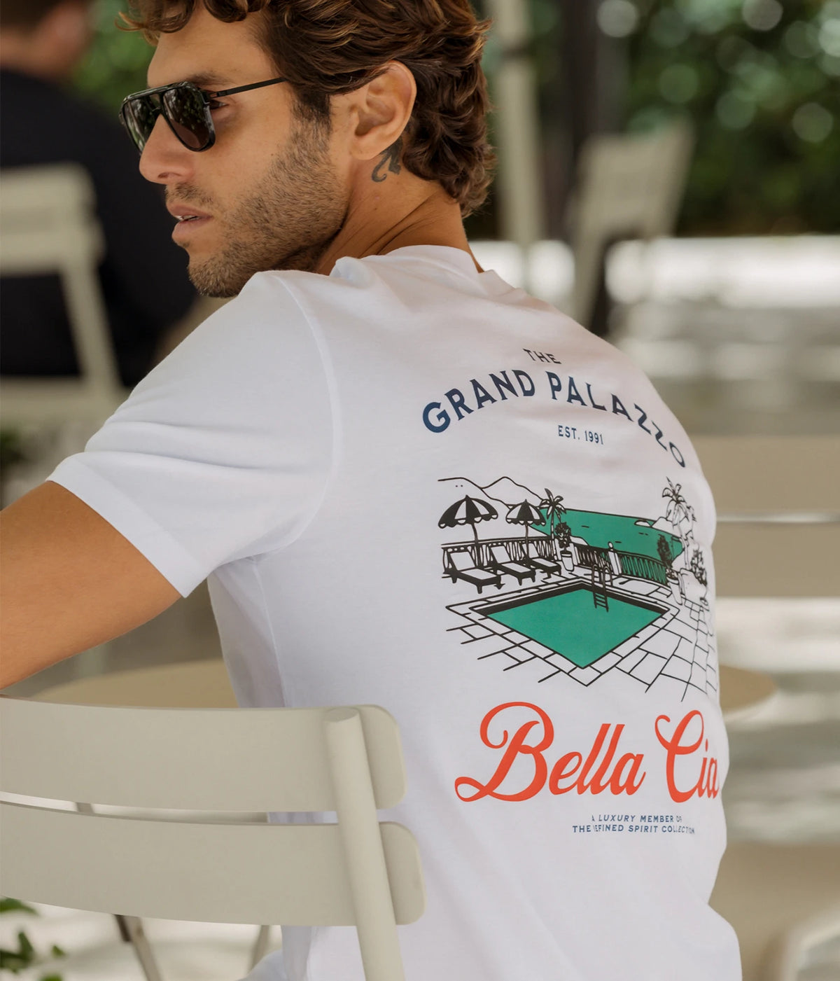 Man wearing The Refined Spirit Grand Palazzo Tee in White with sunglasses, seated outdoors, back view showing the back print