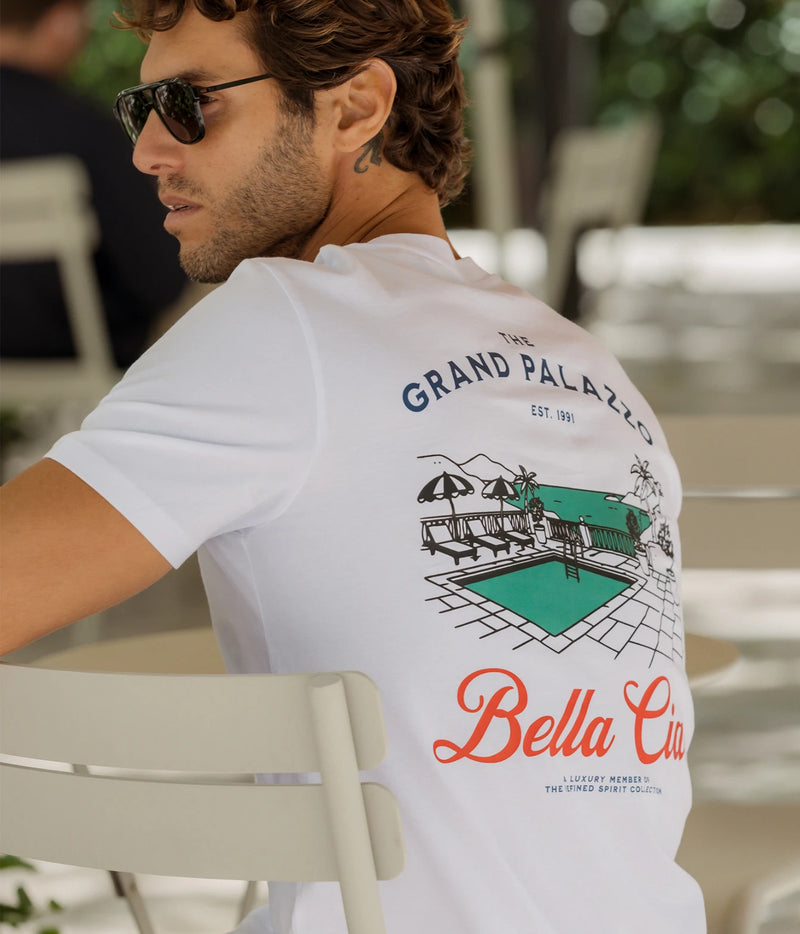 Man wearing The Refined Spirit Grand Palazzo Tee in White with sunglasses, seated outdoors, back view showing the back print