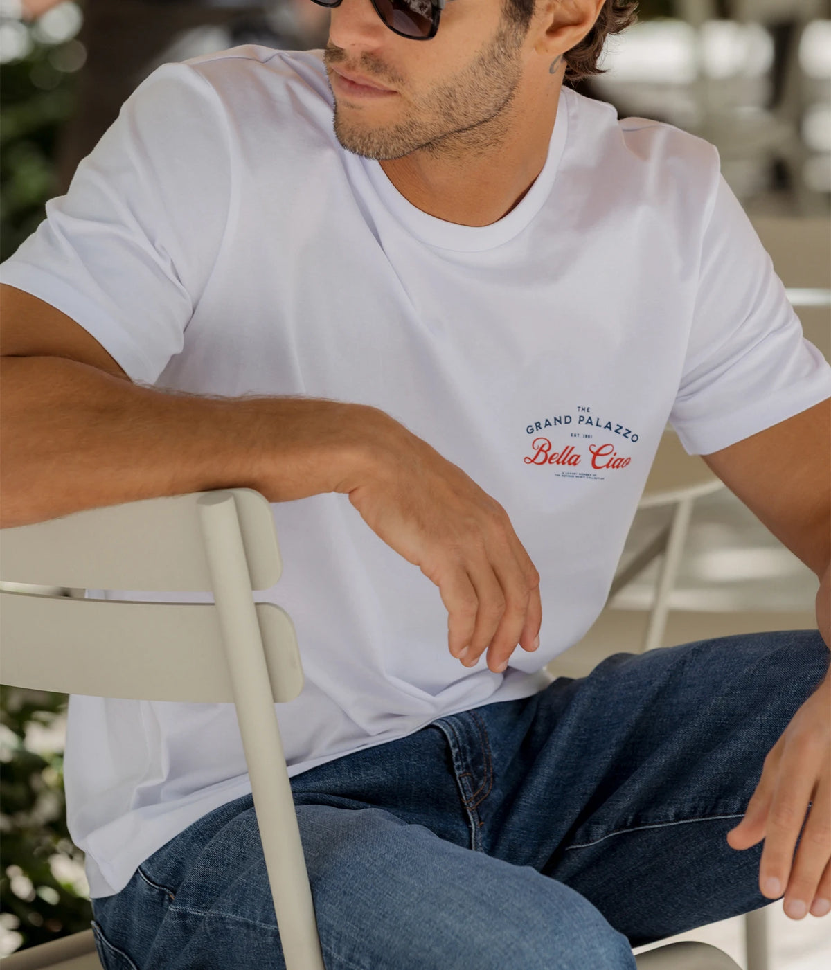 Man wearing The Refined Spirit Grand Palazzo Tee in White with jeans and sunglasses, seated outdoors, front view