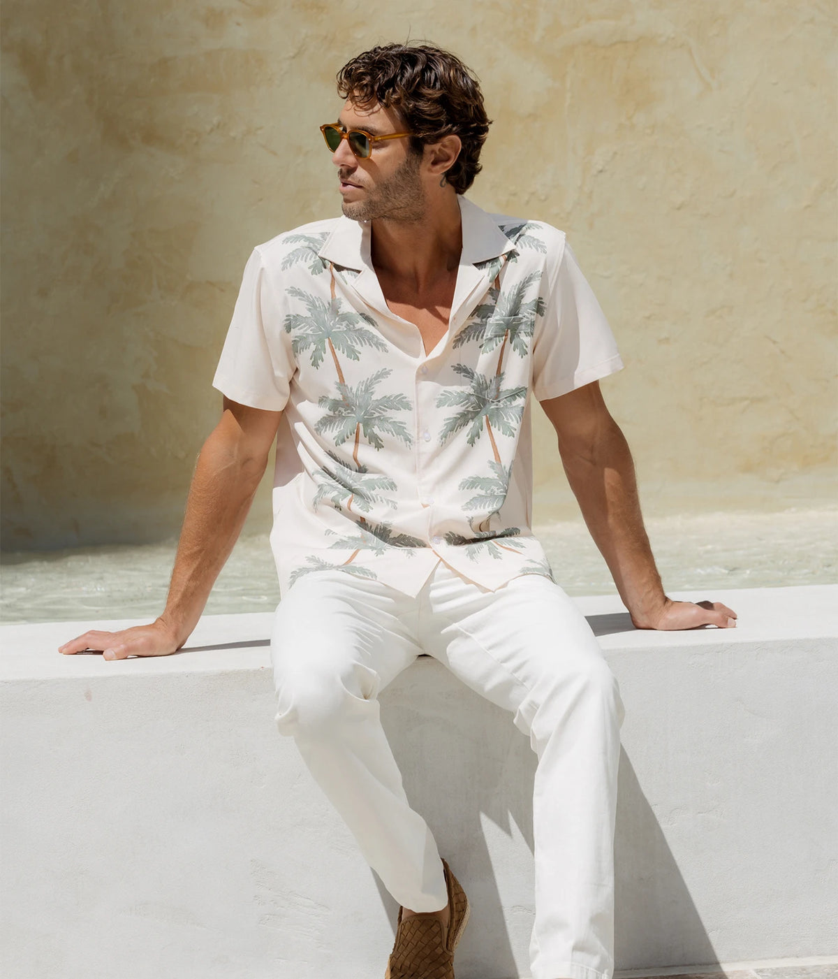 Man wearing Mavrans Palm Row Weekend Shirt in Light Green with white trousers and sunglasses, seated on a ledge, front view