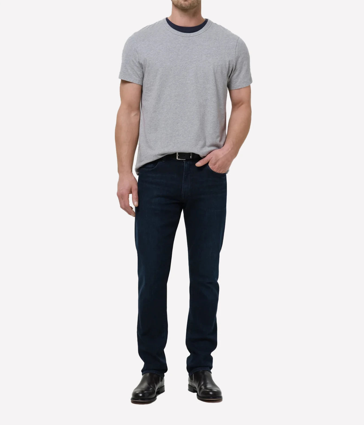 A man wearing a light grey crew-neck t-shirt with a dark navy collar, dark navy blue jeans, a black belt, and black leather dress shoes, standing against a light grey studio background.