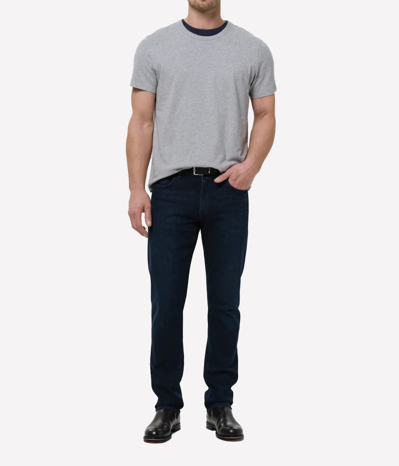 A man wearing a light grey crew-neck t-shirt with a dark navy collar, dark navy blue jeans, a black belt, and black leather dress shoes, standing against a light grey studio background.