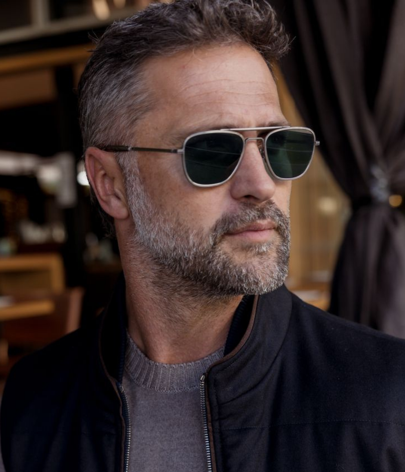 A close-up portrait of a man with a graying beard wearing silver-framed aviator sunglasses with dark green lenses, set against a softly blurred indoor background.
