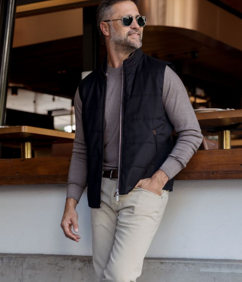 A man wearing a black zip-front puffer vest over a taupe knit sweater, beige trousers, and sunglasses, leaning against a white wall in a modern interior setting.