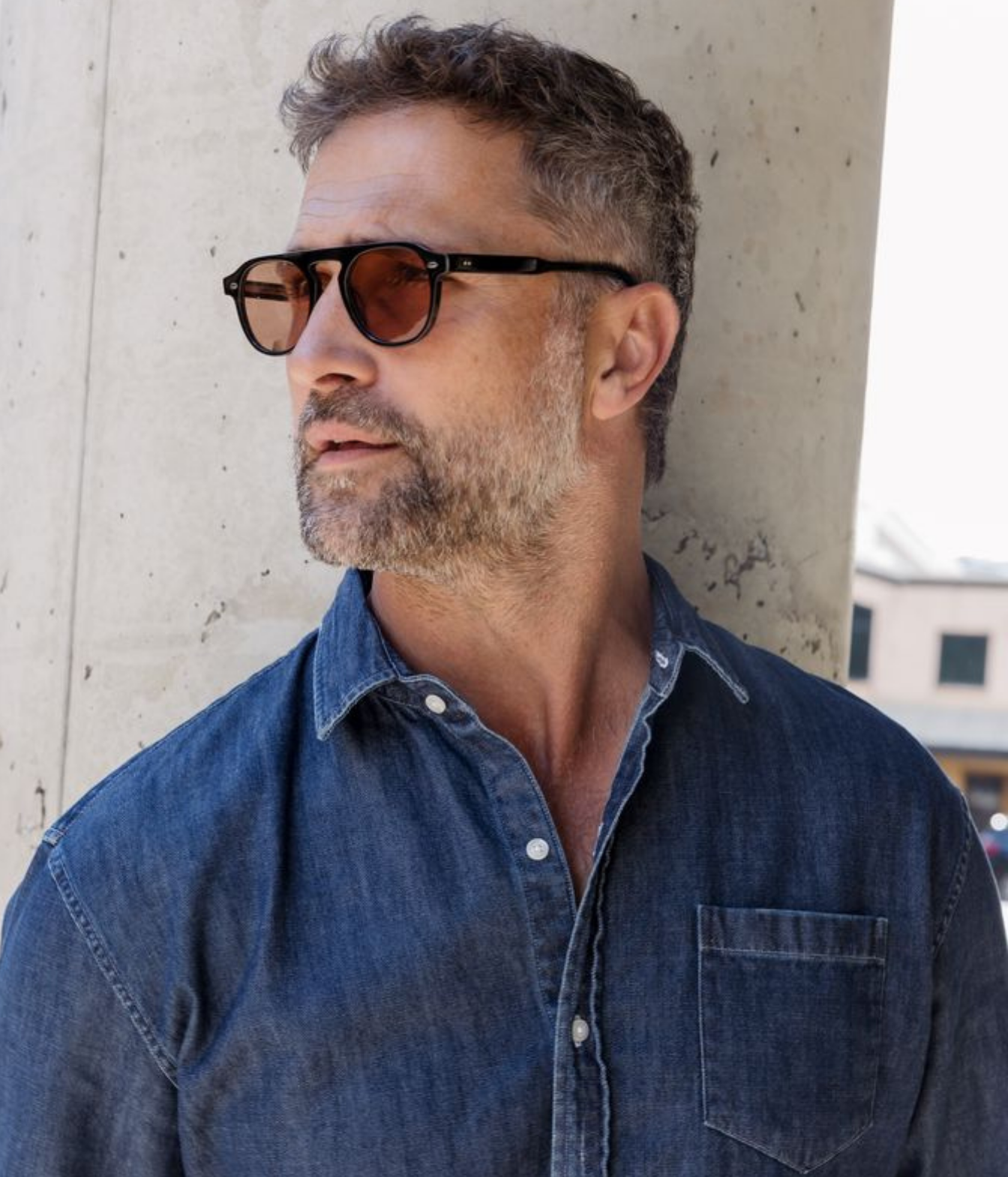 A man with a graying beard and short hair wearing black square sunglasses with brown lenses and a blue denim button-up shirt, leaning against a concrete pillar.