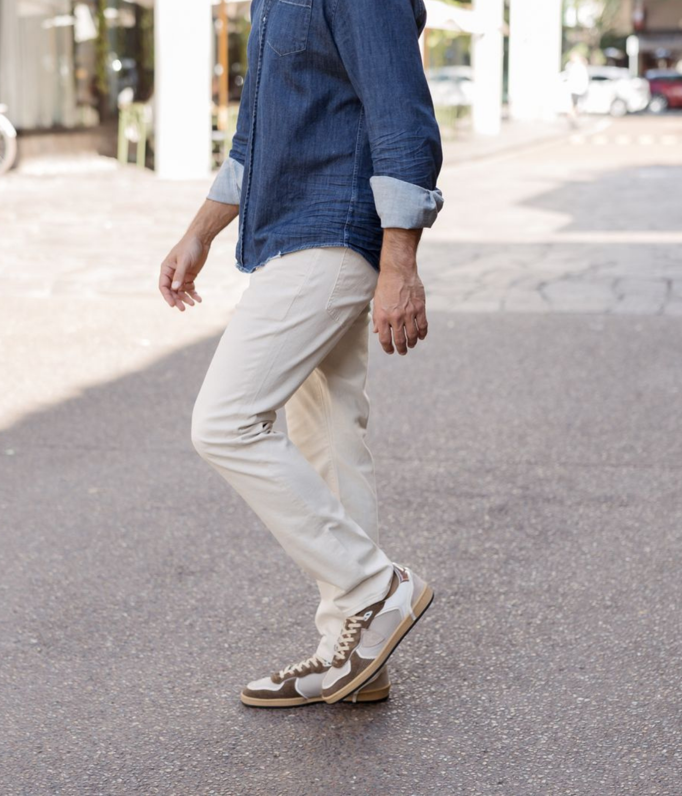 A person walking outdoors on a paved street, wearing off-white chinos, a blue denim shirt, and brown and white sneakers.