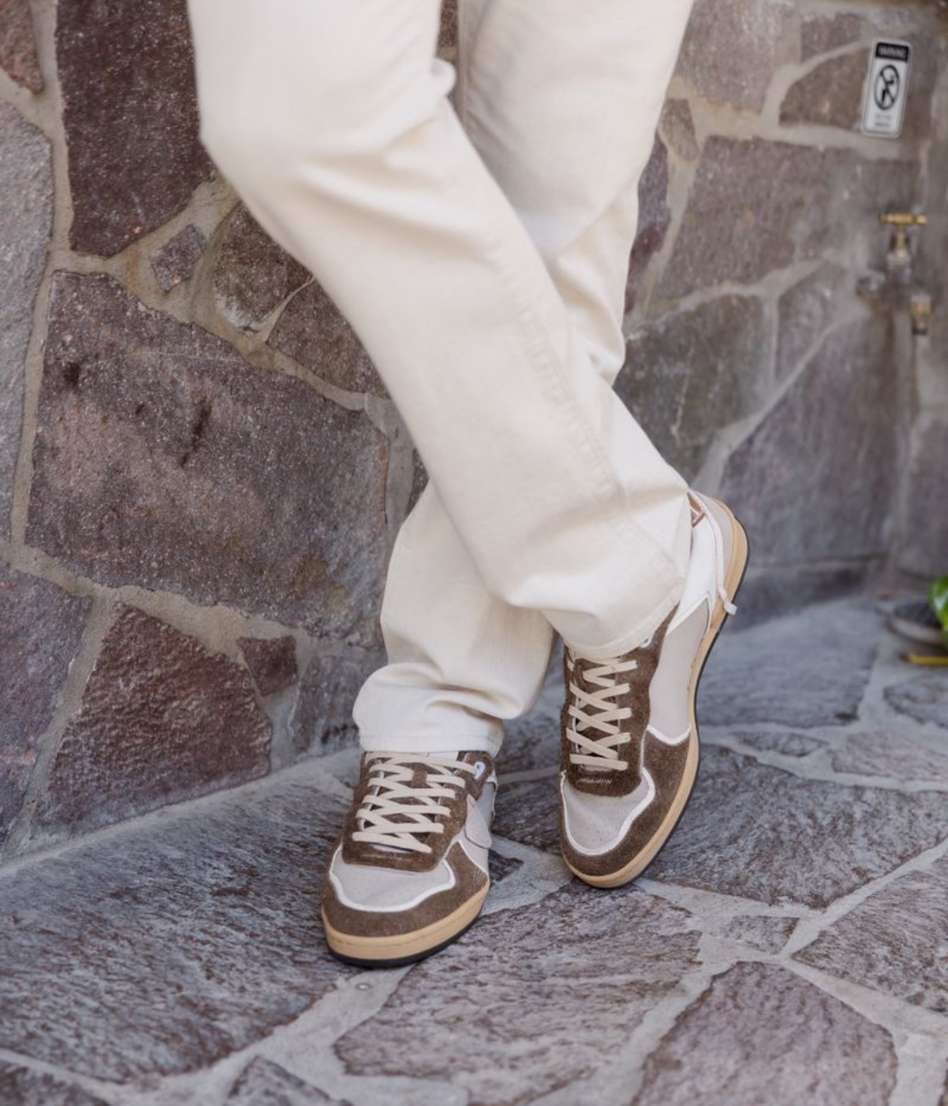 A person wearing off-white pants and brown and beige low-top sneakers, standing on a stone paved floor against a stone wall.