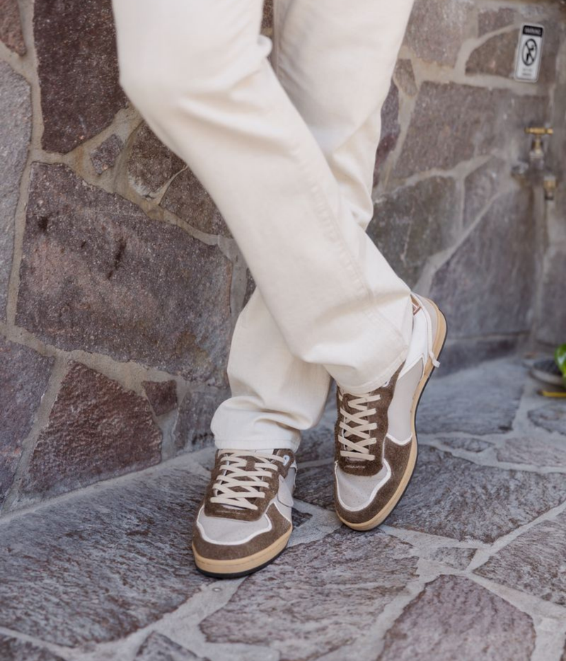 A person wearing off-white pants and brown and beige low-top sneakers, standing on a stone paved floor against a stone wall.