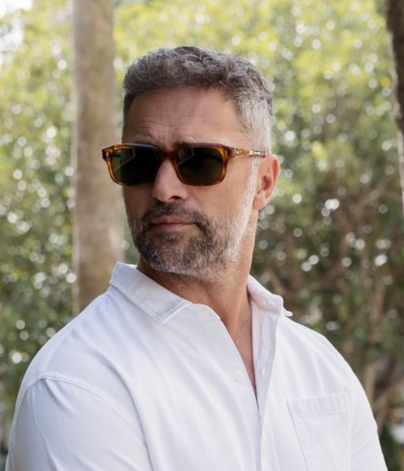 A man with short gray hair and a beard wearing tortoiseshell sunglasses and a white button-up shirt, photographed outdoors with a blurred green and brown background.