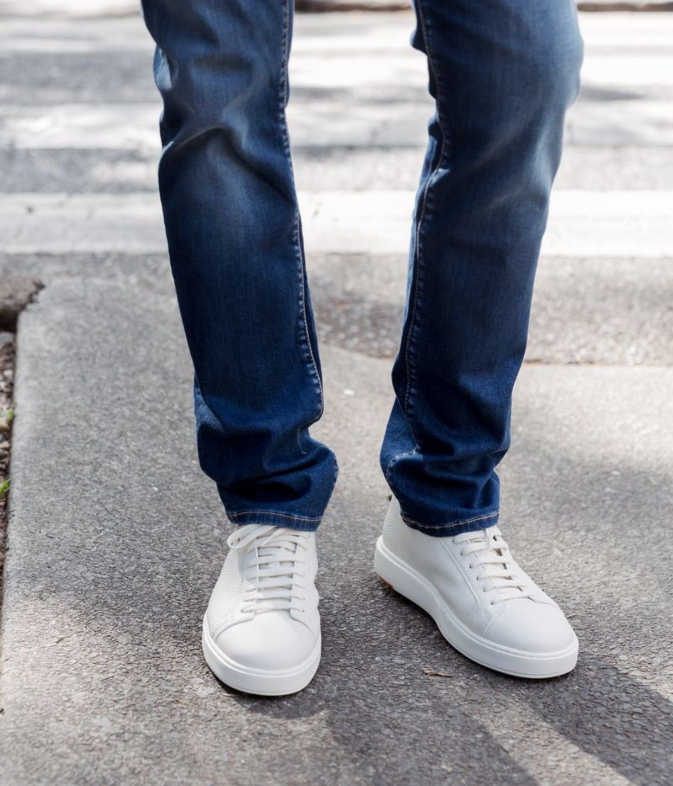 A person wearing dark blue jeans and white low-top sneakers standing on a gray concrete sidewalk outdoors.