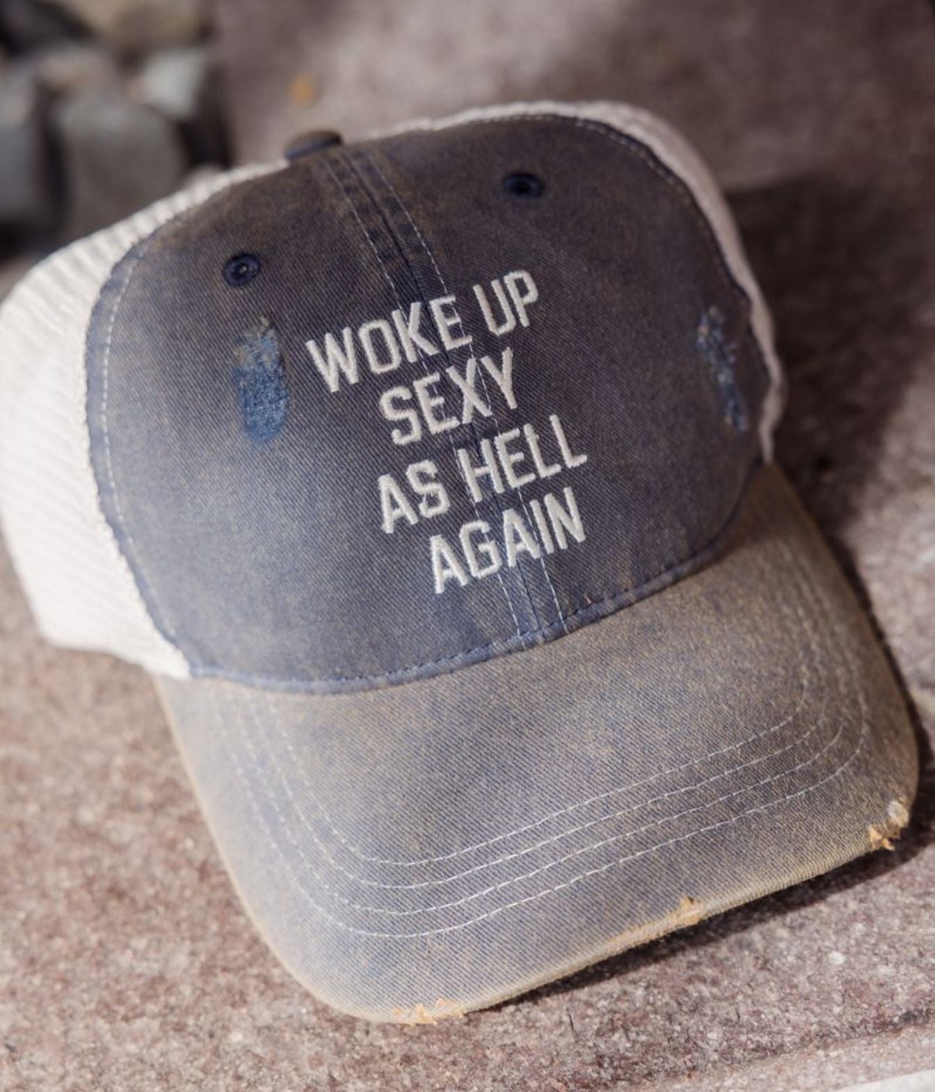 A distressed trucker-style baseball cap with a dark gray front panel and white mesh back, featuring the text 'WOKE UP SEXY AS HELL AGAIN' in white on the front.
