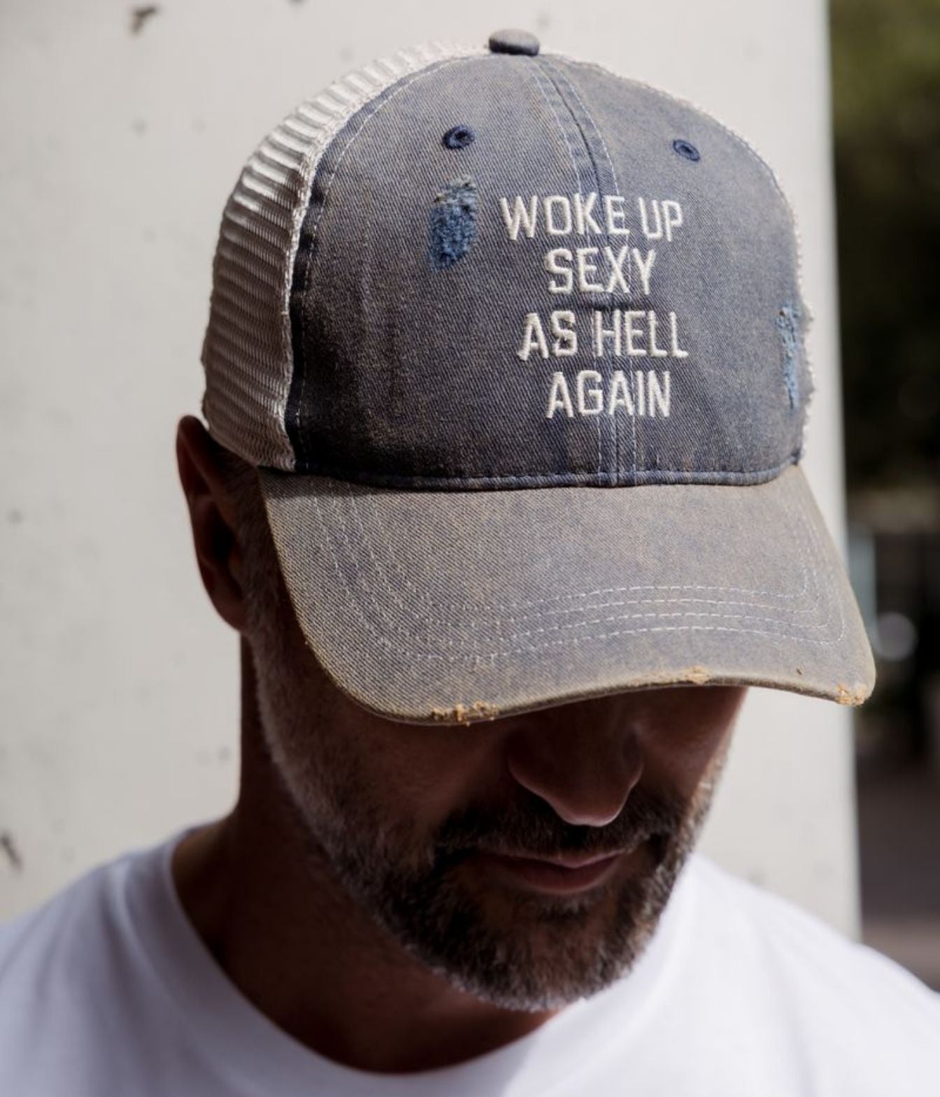 A man wearing a distressed trucker hat with a gray washed front panel and brim, and a beige mesh back. The front of the hat features white embroidered text that reads 'WOKE UP SEXY AS HELL AGAIN'.