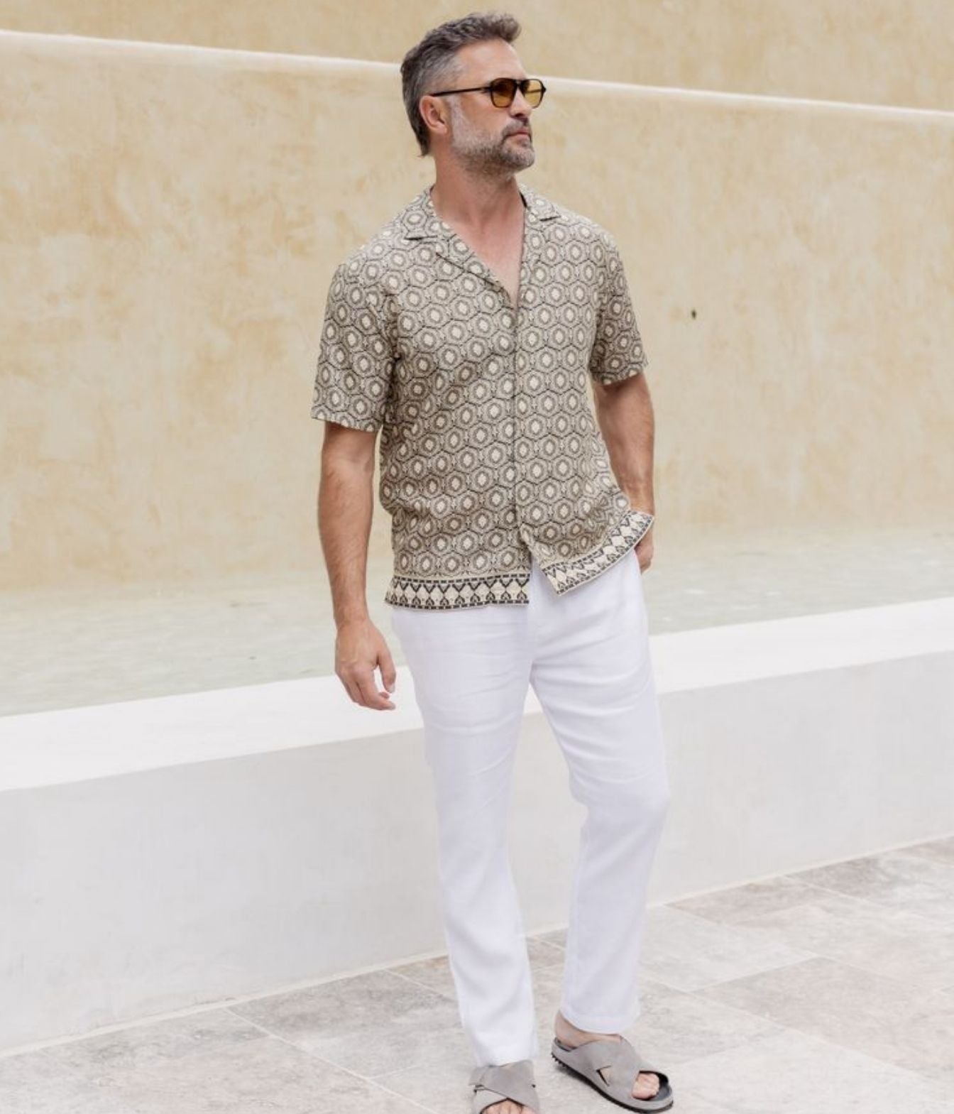 A man wearing a short-sleeve button-up shirt with a beige and brown geometric pattern, white trousers, and sunglasses, standing in front of a beige stucco wall.