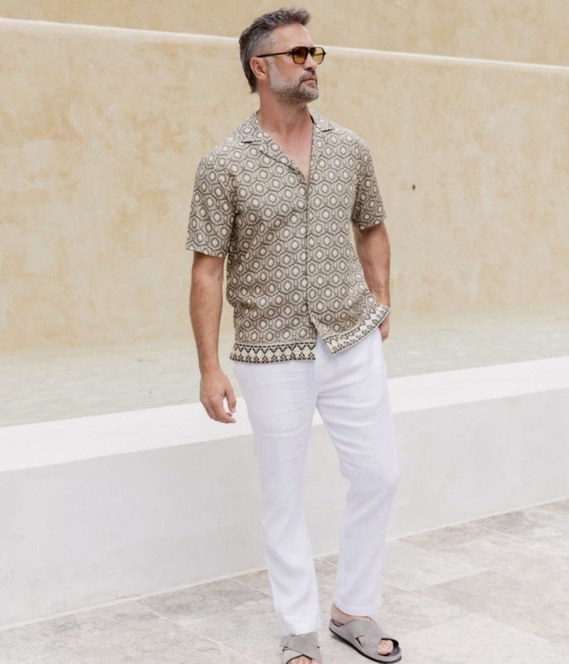 A man wearing a short-sleeve button-up shirt with a beige and brown geometric pattern, white trousers, and sunglasses, standing in front of a beige stucco wall.