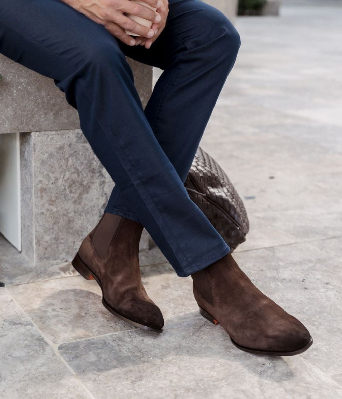 A close-up lifestyle shot of a person sitting on a stone bench, wearing dark brown suede Chelsea boots with elastic side panels and navy blue trousers.