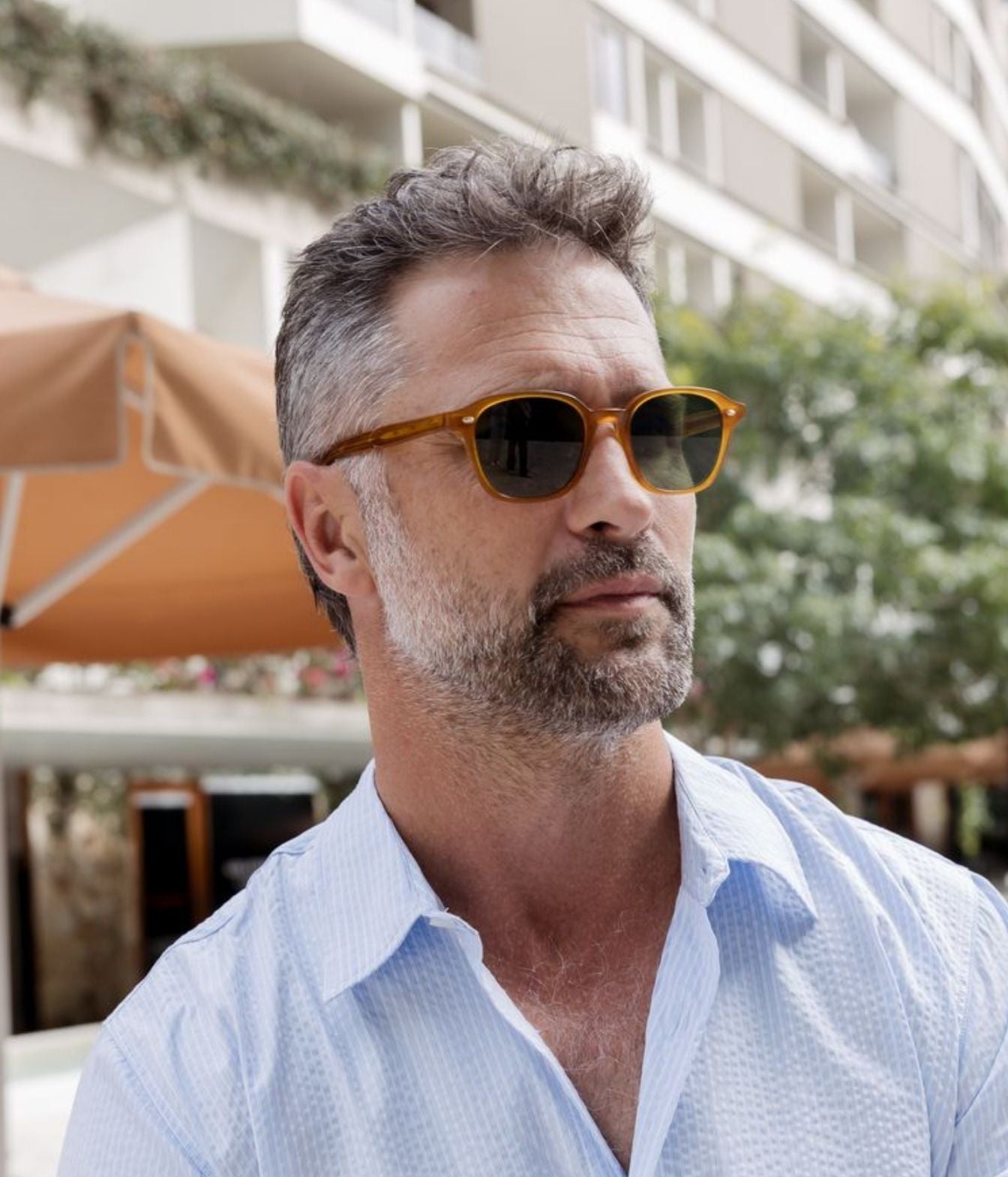 A man wearing amber acetate round-frame sunglasses with dark lenses and a light blue textured button-down shirt, photographed outdoors with a blurred background of buildings and patio umbrellas.