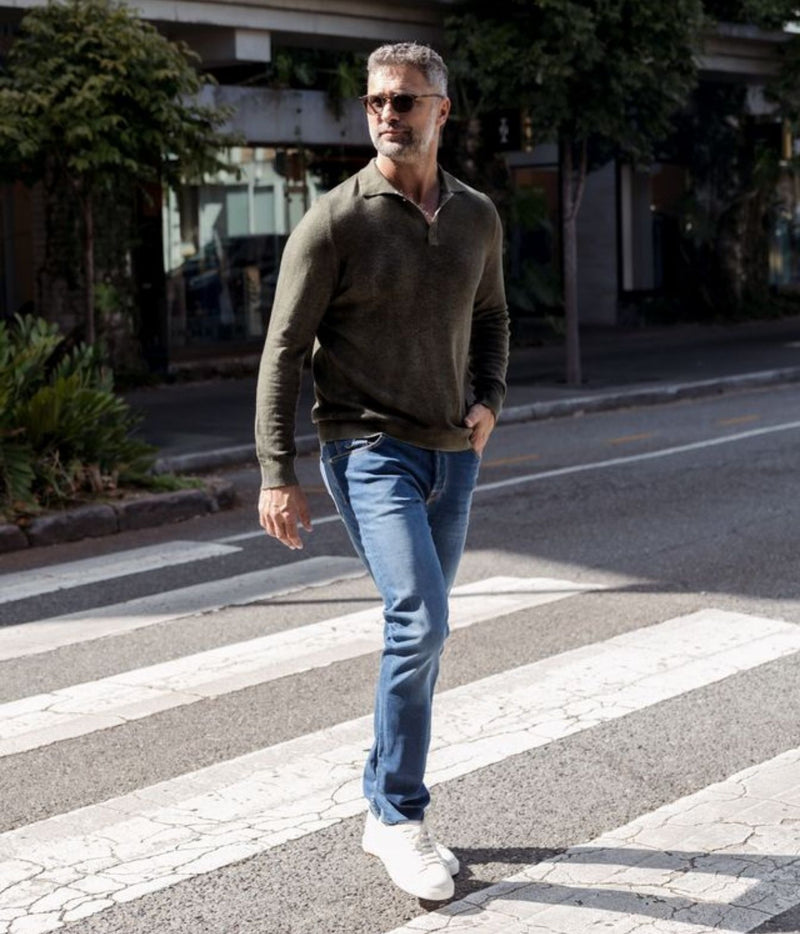 A man walking across a city crosswalk wearing an olive green long-sleeve polo sweater, blue jeans, and white sneakers.