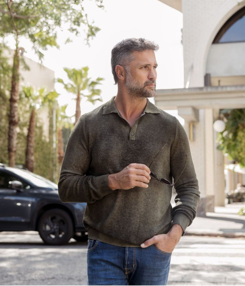 A man wearing a dark olive green long-sleeve polo shirt and blue jeans, standing outdoors in front of a building and palm trees.