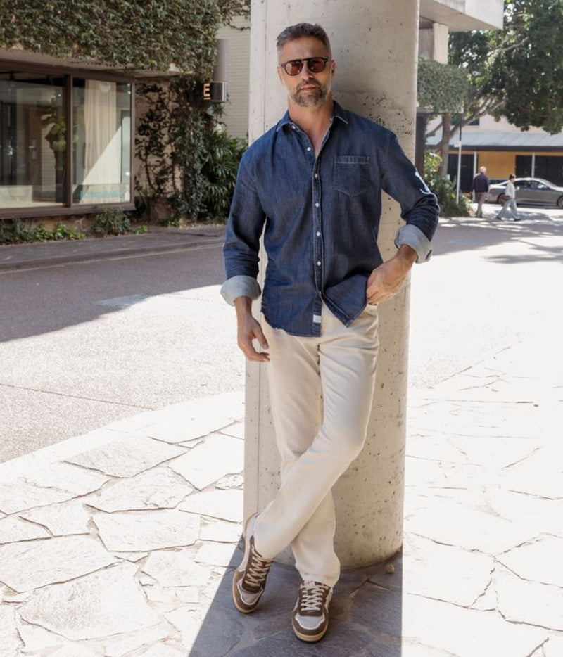 A man wearing a dark blue denim button-up shirt, beige trousers, and brown sneakers leans against a concrete pillar on a sunny outdoor street.