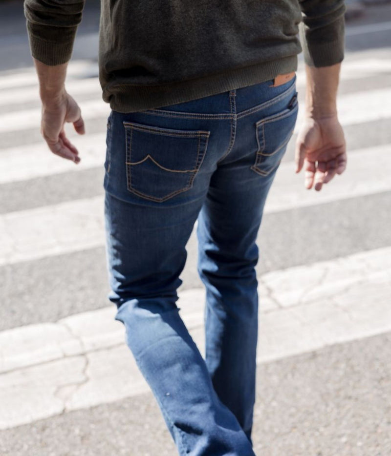 A person viewed from behind wearing blue jeans with contrast tan stitching, walking across a striped crosswalk outdoors.