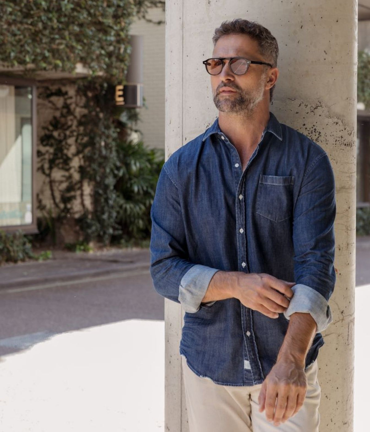 A man wearing a dark blue chambray button-down shirt and beige pants leans against a concrete pillar in an outdoor setting.