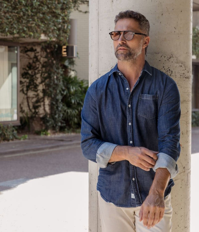 A man wearing a dark blue chambray button-down shirt and beige pants leans against a concrete pillar in an outdoor setting.