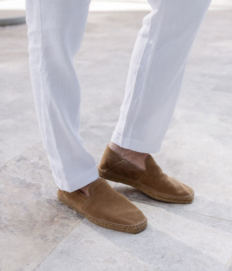 A person wearing white trousers and tan suede slip-on espadrille shoes on a light stone floor.