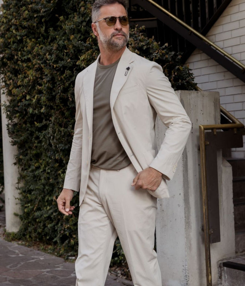 A man wearing a light beige two-piece suit with a taupe t-shirt and sunglasses, standing outdoors next to a concrete pillar and green ivy-covered wall.